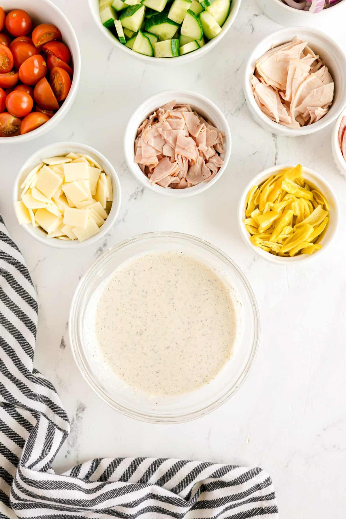A bowl of creamy dressing surrounded by small bowls containing sliced cucumbers, cherry tomatoes, deli meat, cheese slices, and artichoke hearts on a white surface with a striped towel.