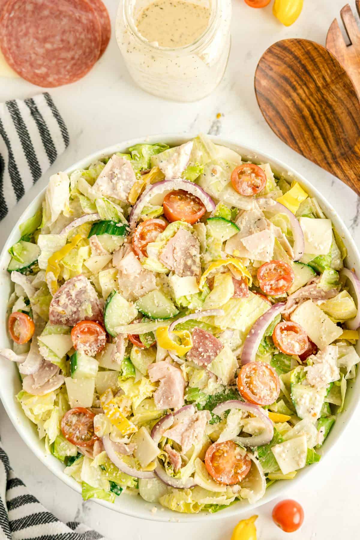 A large bowl of chopped salad with lettuce, tomatoes, cucumbers, red onions, cheese, salami, and dressing, surrounded by salad utensils and additional ingredients.
