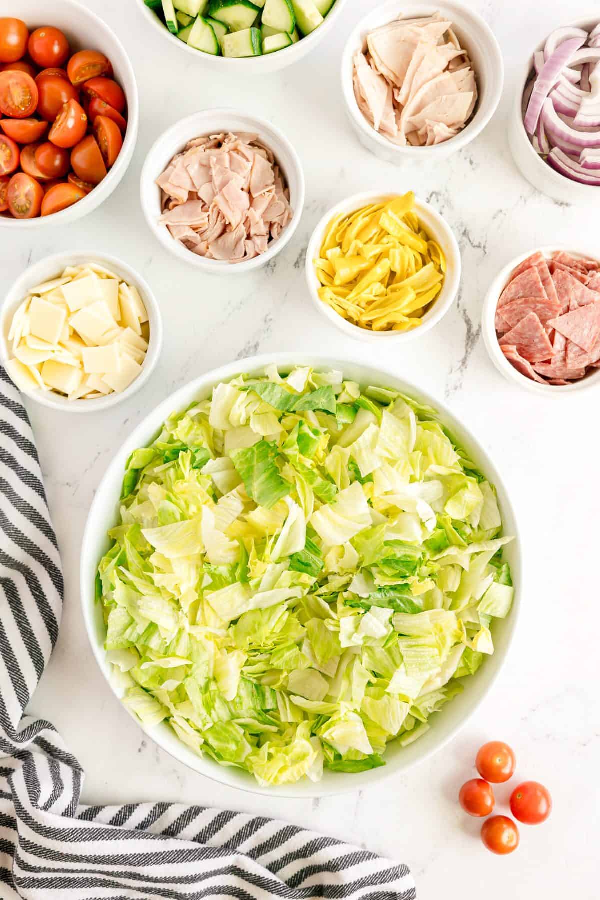 A bowl of chopped lettuce surrounded by small bowls of sliced cucumber, cherry tomatoes, lunch meats, cheese, and red onion on a marble surface.