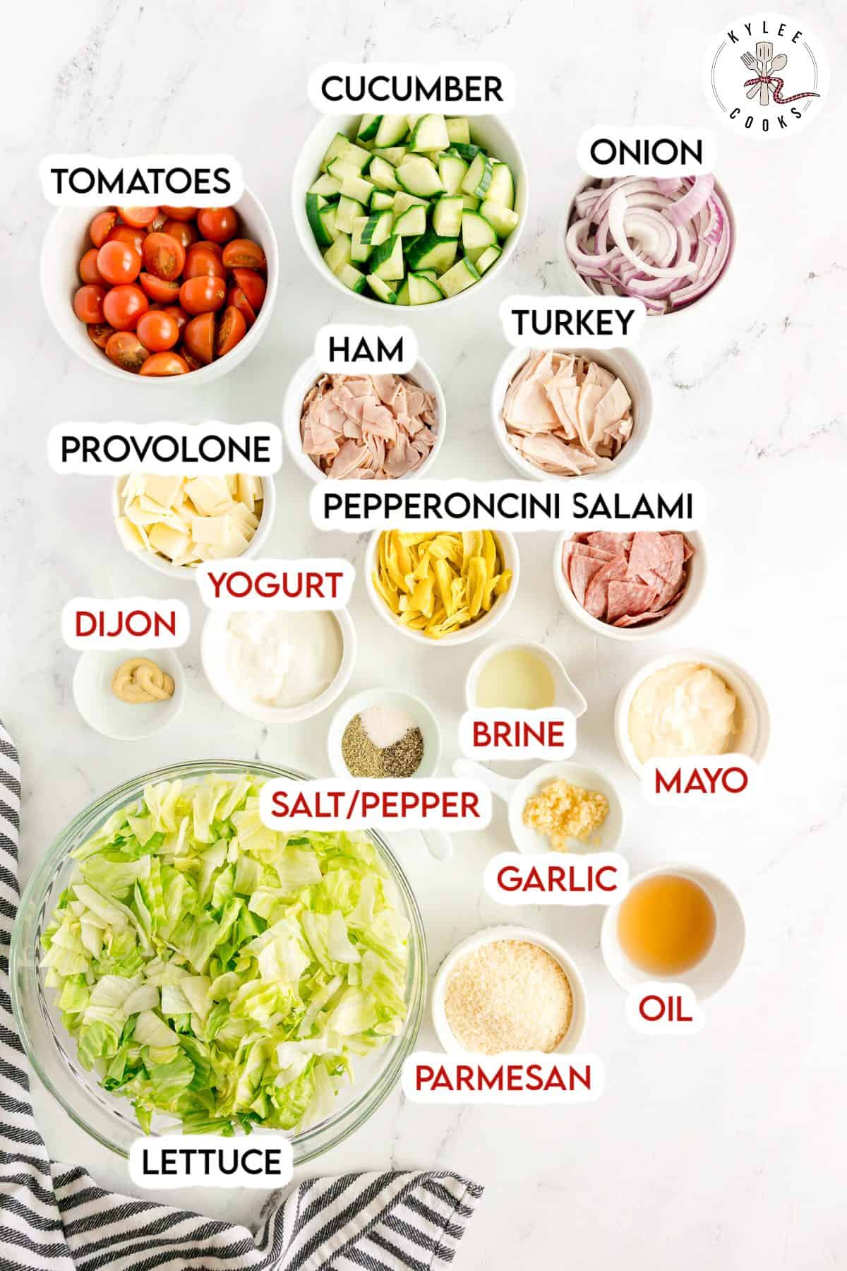 Top-down view of salad ingredients in separate bowls, including lettuce, tomato, cucumber, onion, turkey, ham, salami, provolone, pepperoncini, parmesan, and various dressings and seasonings.