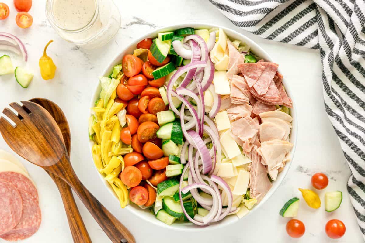 A bowl of salad with sliced red onions, cucumbers, cherry tomatoes, lettuce, cheese, salami, and ham, next to wooden utensils and a jar of dressing on a white surface.