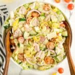 A bowl of chopped salad with lettuce, cherry tomatoes, cucumber, red onion, banana peppers, sliced meats, cheese, and two wooden serving spoons.