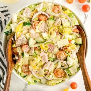 A bowl of chopped salad with lettuce, cherry tomatoes, cucumber, red onion, banana peppers, sliced meats, cheese, and two wooden serving spoons.