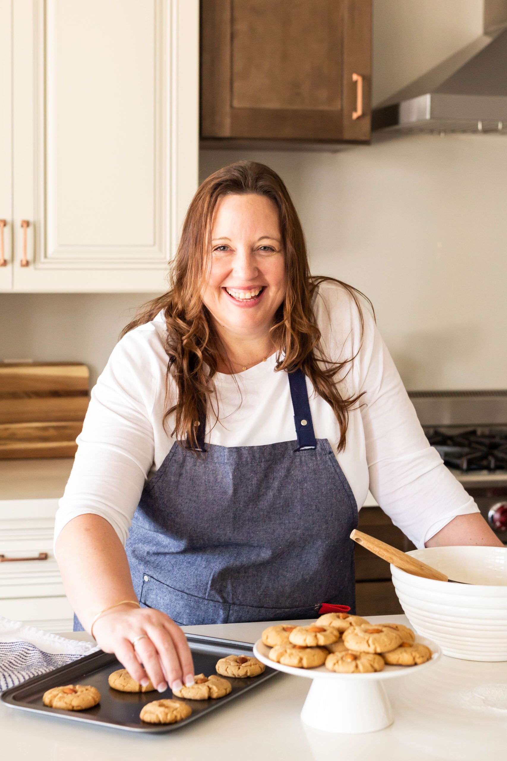 Woman wearing a blue apron smiles while placing freshly baked cookies on a tray in a bright kitchen.