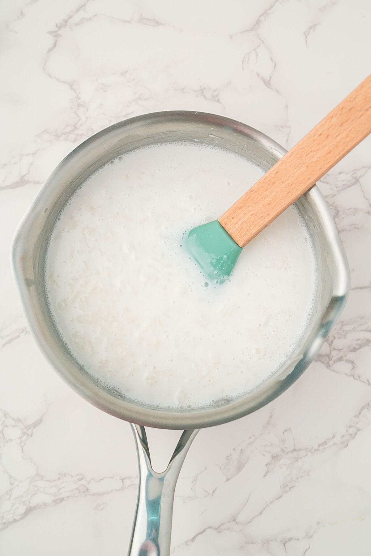 A saucepan filled with rice and milk being stirred with a wooden spatula on a marble countertop.