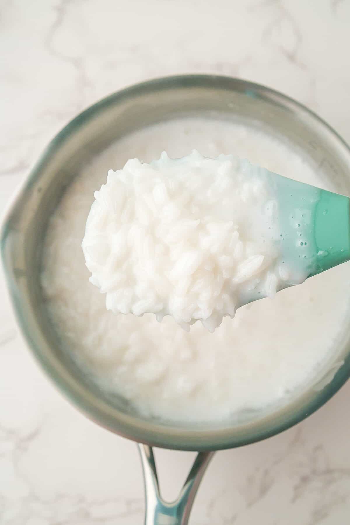 A close-up of a spoonful of cooked rice and milk held above a saucepan filled with the same mixture on a light countertop.