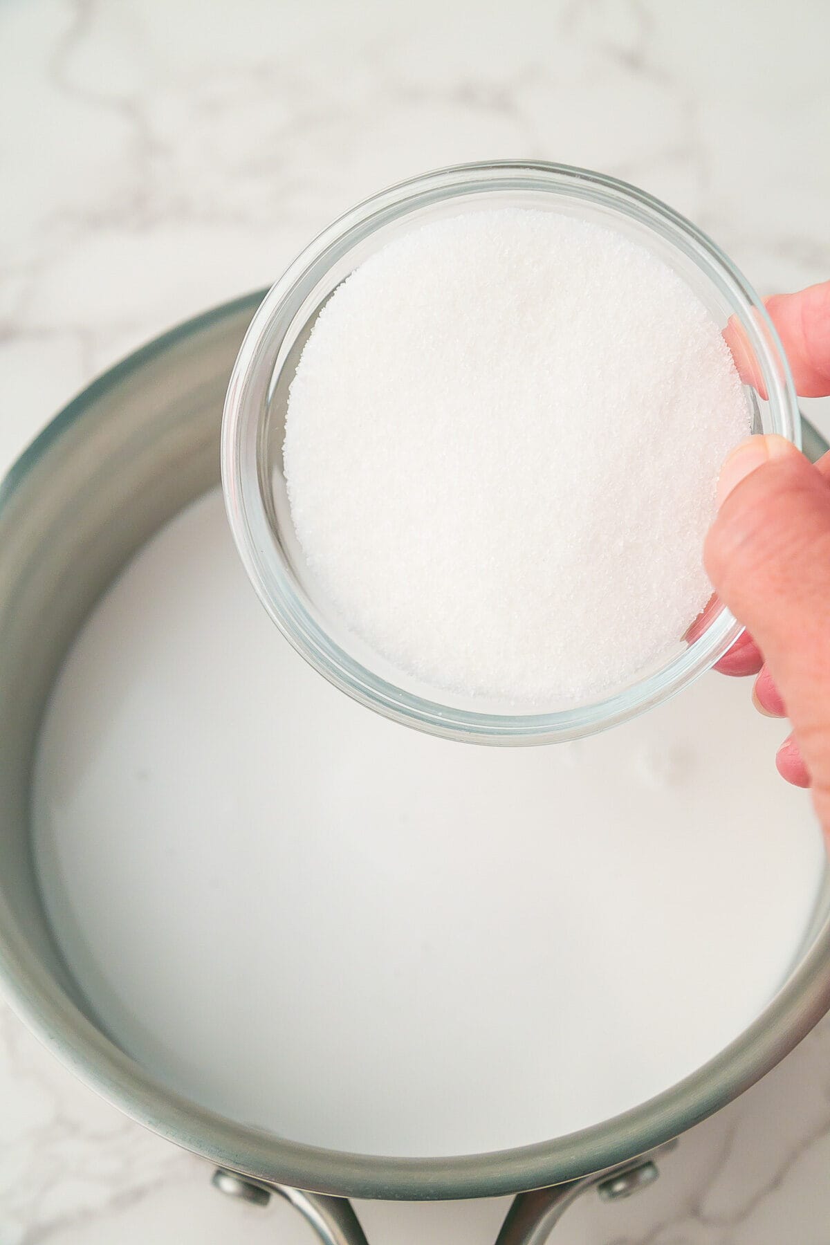 A hand holds a small bowl of granulated sugar above a saucepan filled with milk on a marble countertop.
