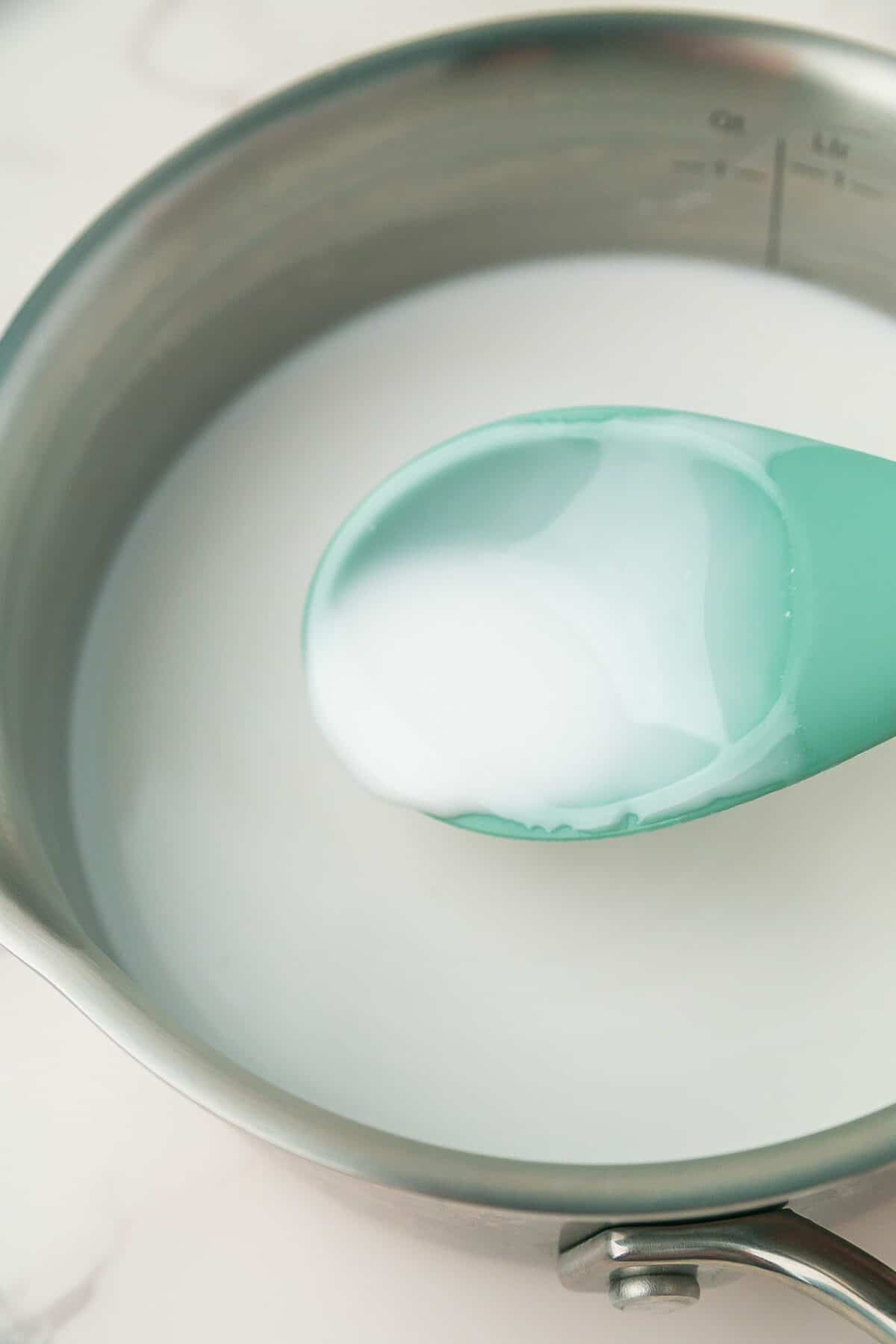 A saucepan filled with a white liquid, possibly thickened, being stirred with a teal silicone spoon.