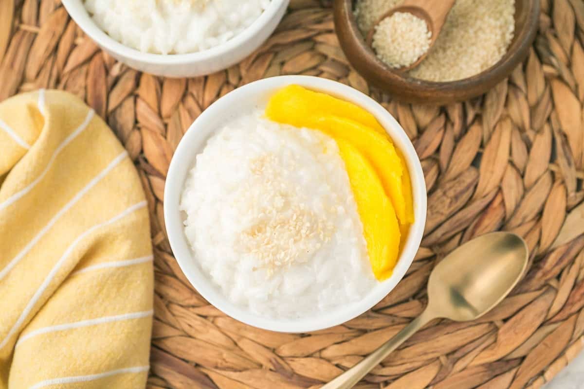 A bowl of mango sticky rice topped with sesame seeds and fresh mango slices sits on a woven mat beside a spoon, yellow striped napkin, and a small bowl of sesame seeds.