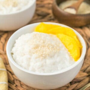 A white bowl of mango sticky rice topped with sesame seeds, served with slices of mango on the side, placed on a woven mat.