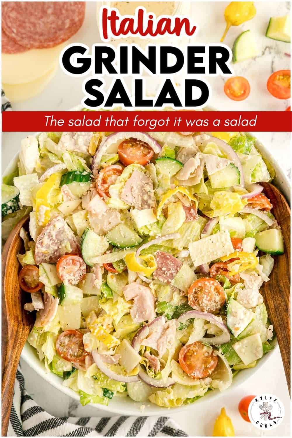A bowl of Italian grinder salad with lettuce, sliced meats, cheese, tomatoes, cucumbers, and peppers, mixed and served with wooden utensils.