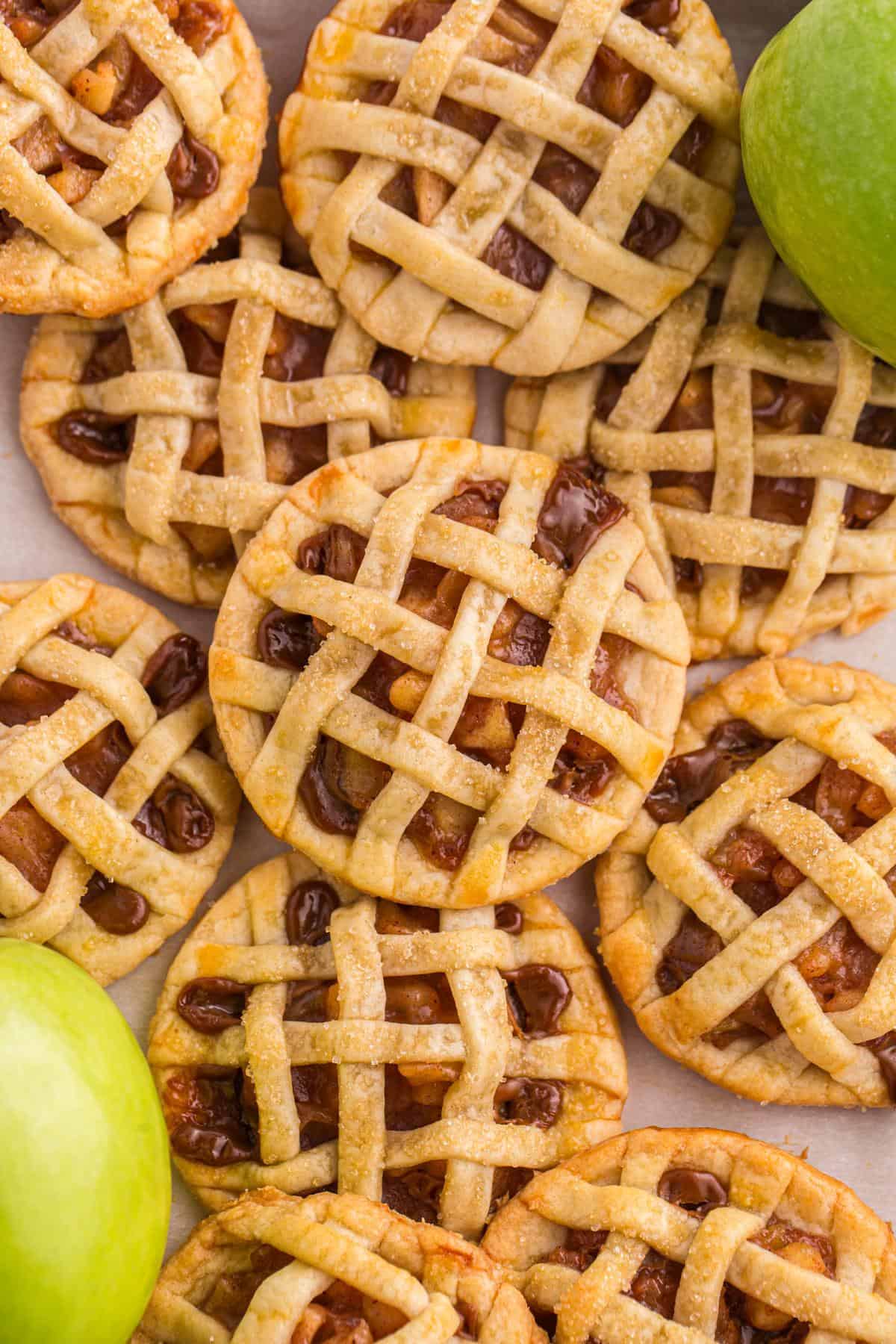 Mini lattice-topped apple pies are arranged closely together, with a few green apples placed next to them.