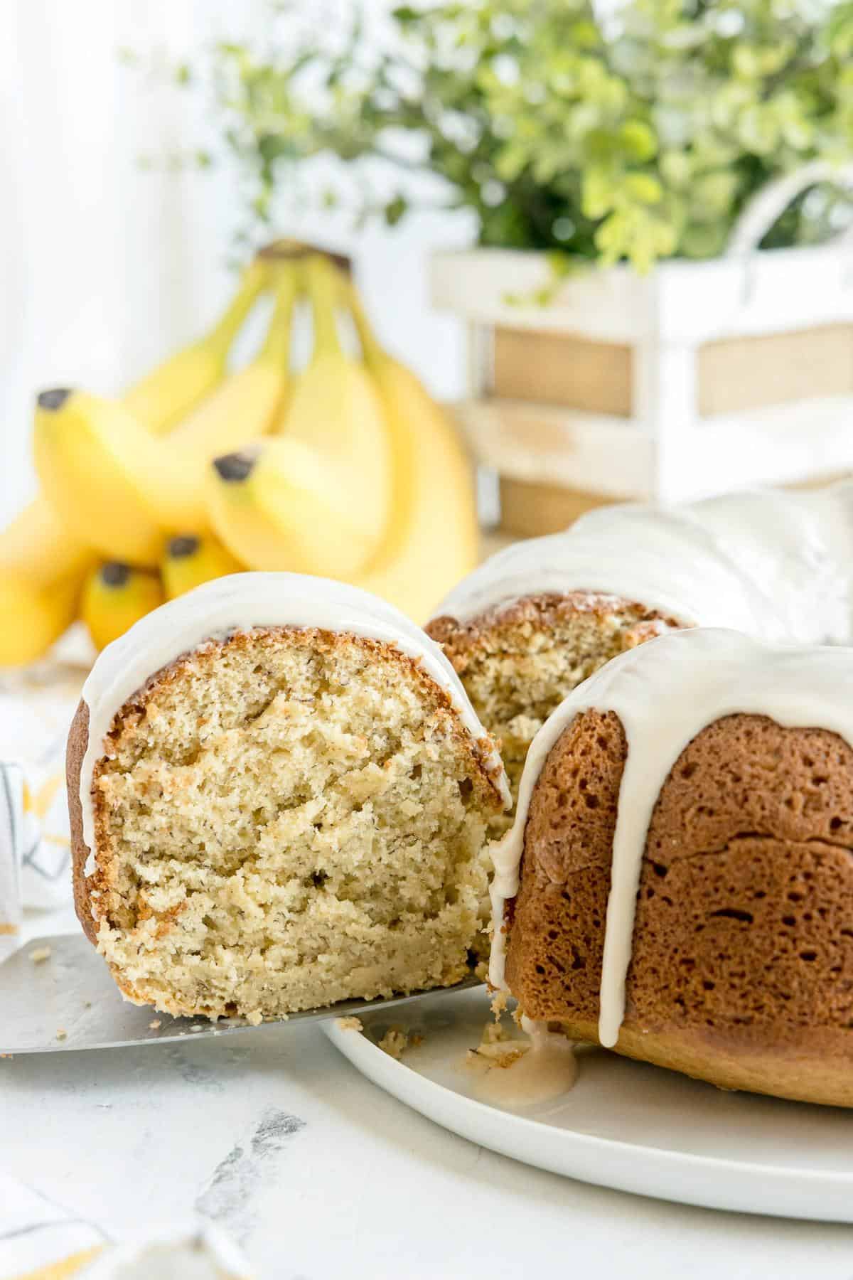 A sliced bundt cake with white icing sits on a plate, with bananas and a decorative plant in the background.