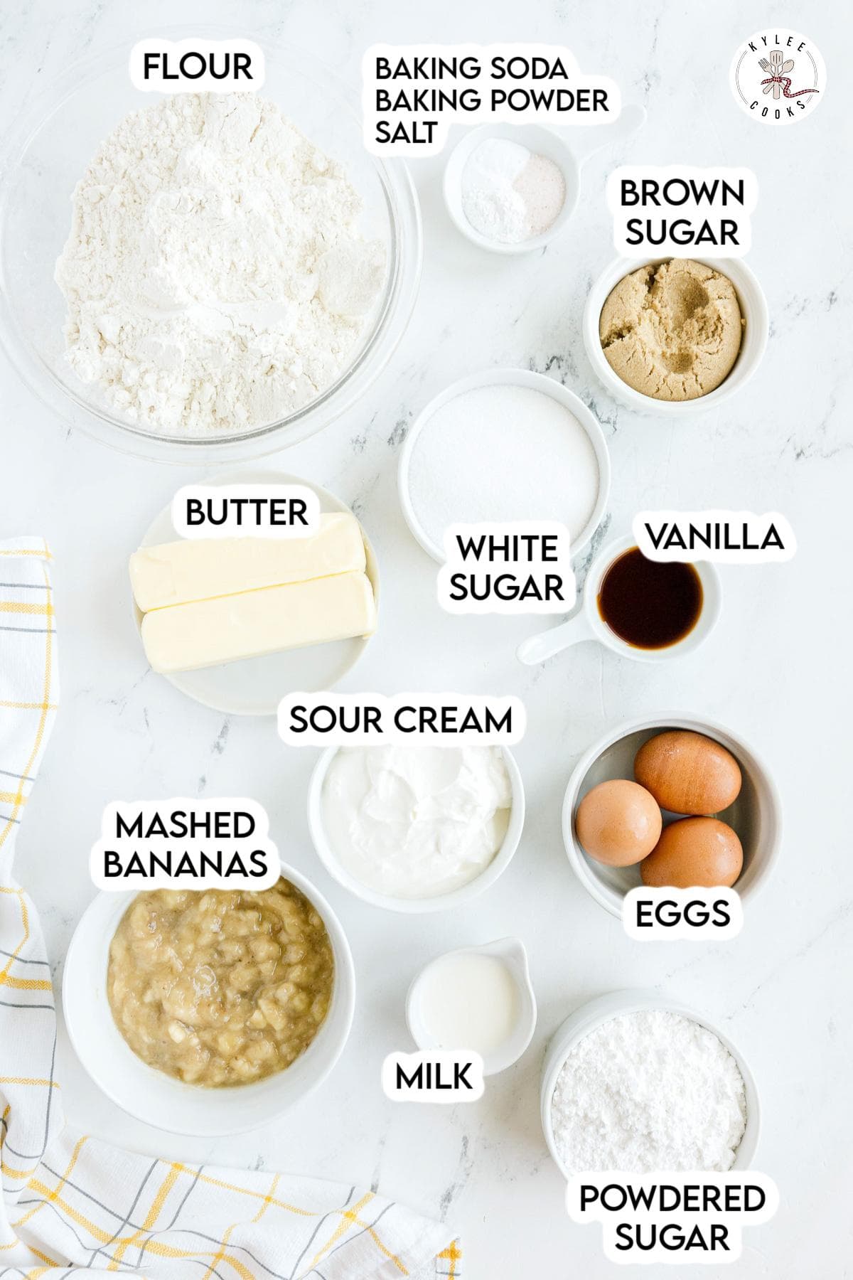Various baking ingredients including flour, brown sugar, white sugar, butter, sour cream, mashed bananas, eggs, milk, vanilla, powdered sugar, and a bowl of baking soda, baking powder, and salt.