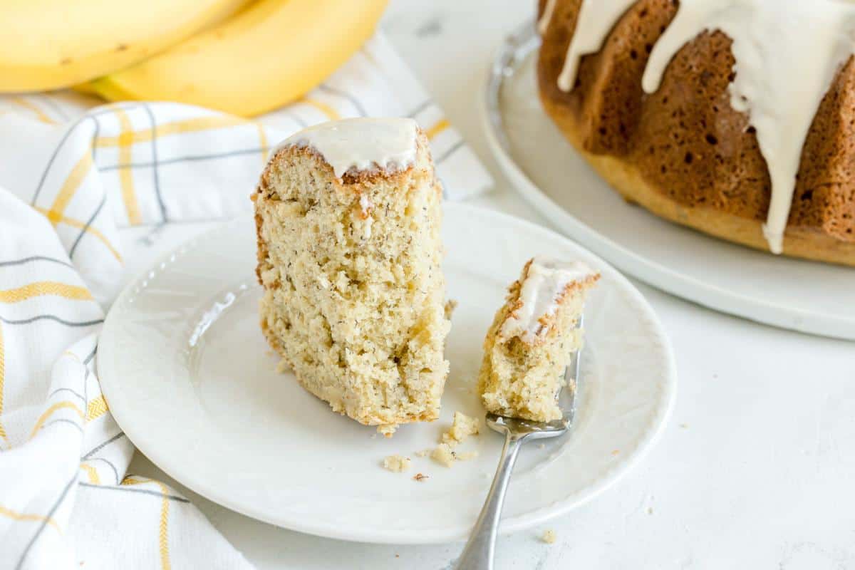 A slice of banana bundt cake with white icing on a plate, with a fork holding a piece, and the rest of the cake and bananas in the background.