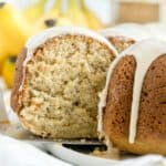 A sliced bundt cake with a light glaze on top, showing a moist, fluffy interior. Bananas are blurred in the background.