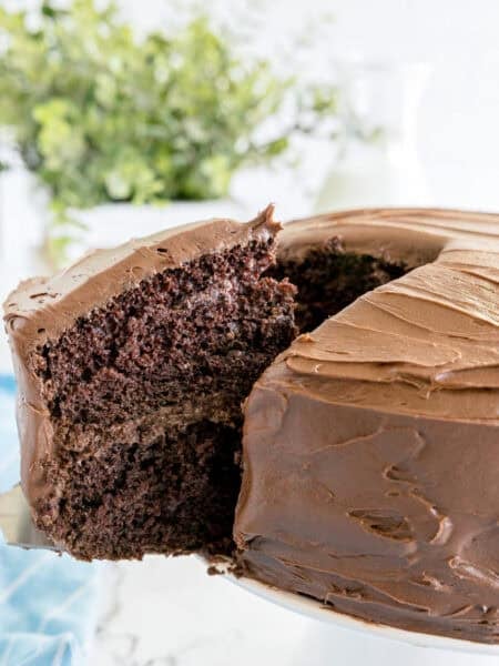 A two-layer chocolate cake with chocolate frosting is being served, with one slice lifted from the whole cake on a white cake stand.