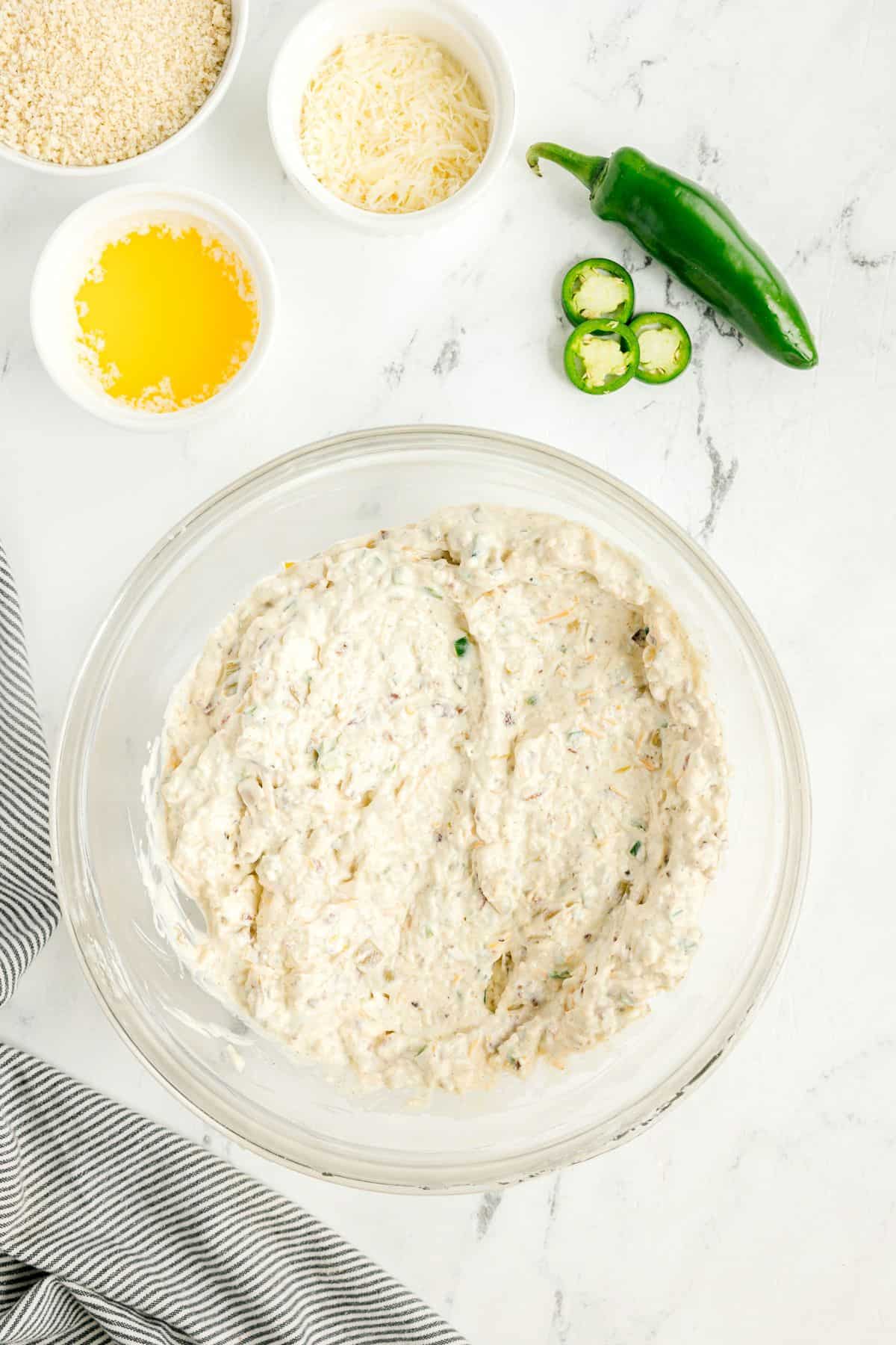 A glass bowl containing a creamy, mixed cheese filling sits on a marble surface beside a sliced jalapeño, melted butter, breadcrumbs, and shredded cheese in small bowls.