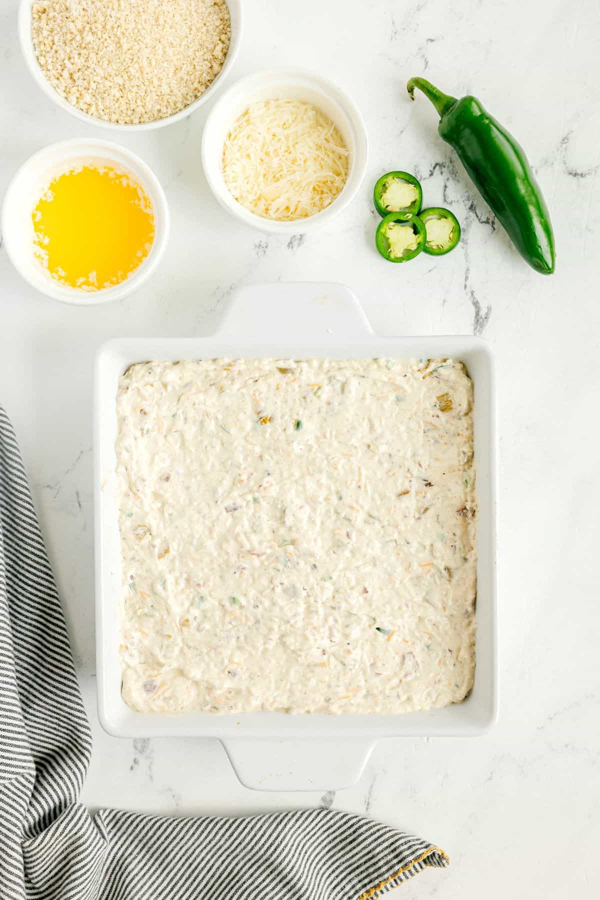 Square baking dish filled with creamy dip mixture, surrounded by bowls of breadcrumbs, melted butter, shredded cheese, and fresh jalapeño on a white surface.