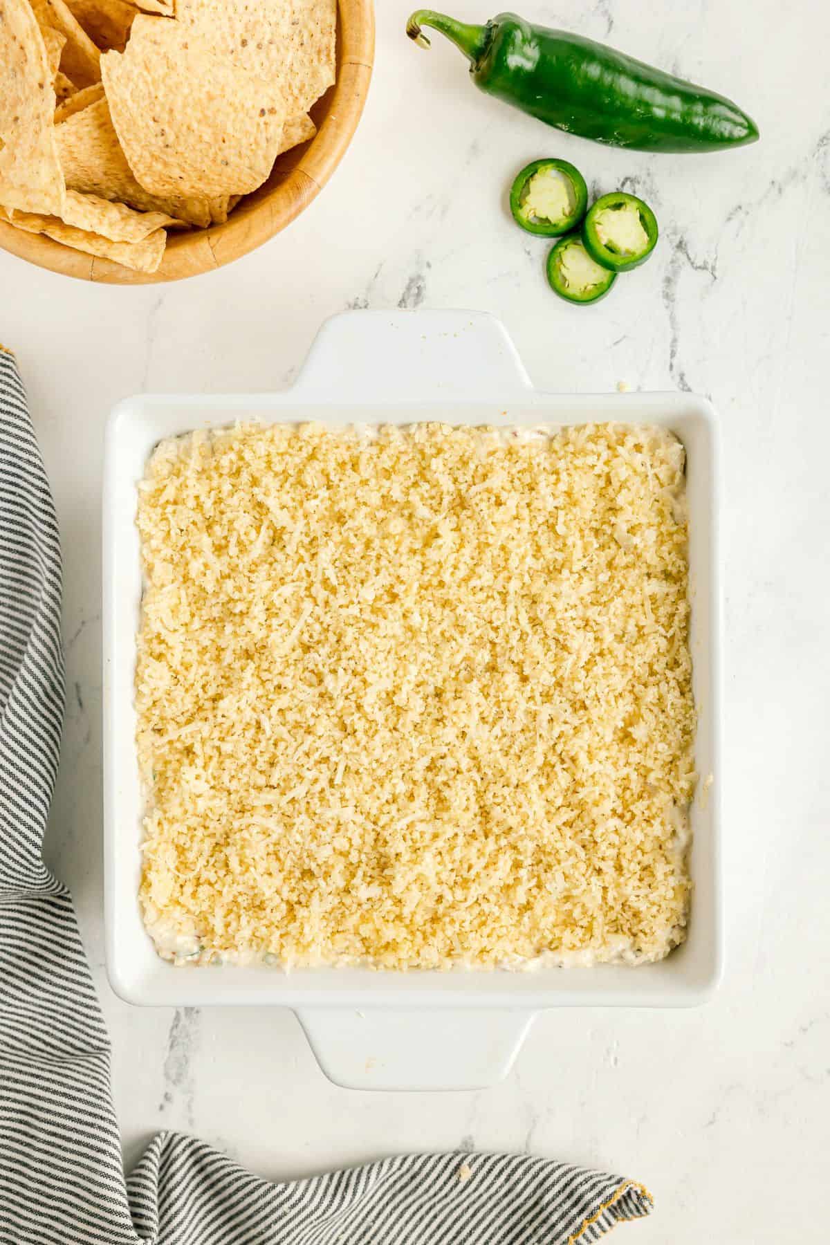 A white baking dish filled with a crumb-topped casserole sits on a marble surface next to tortilla chips, a sliced jalapeño, and a striped cloth.