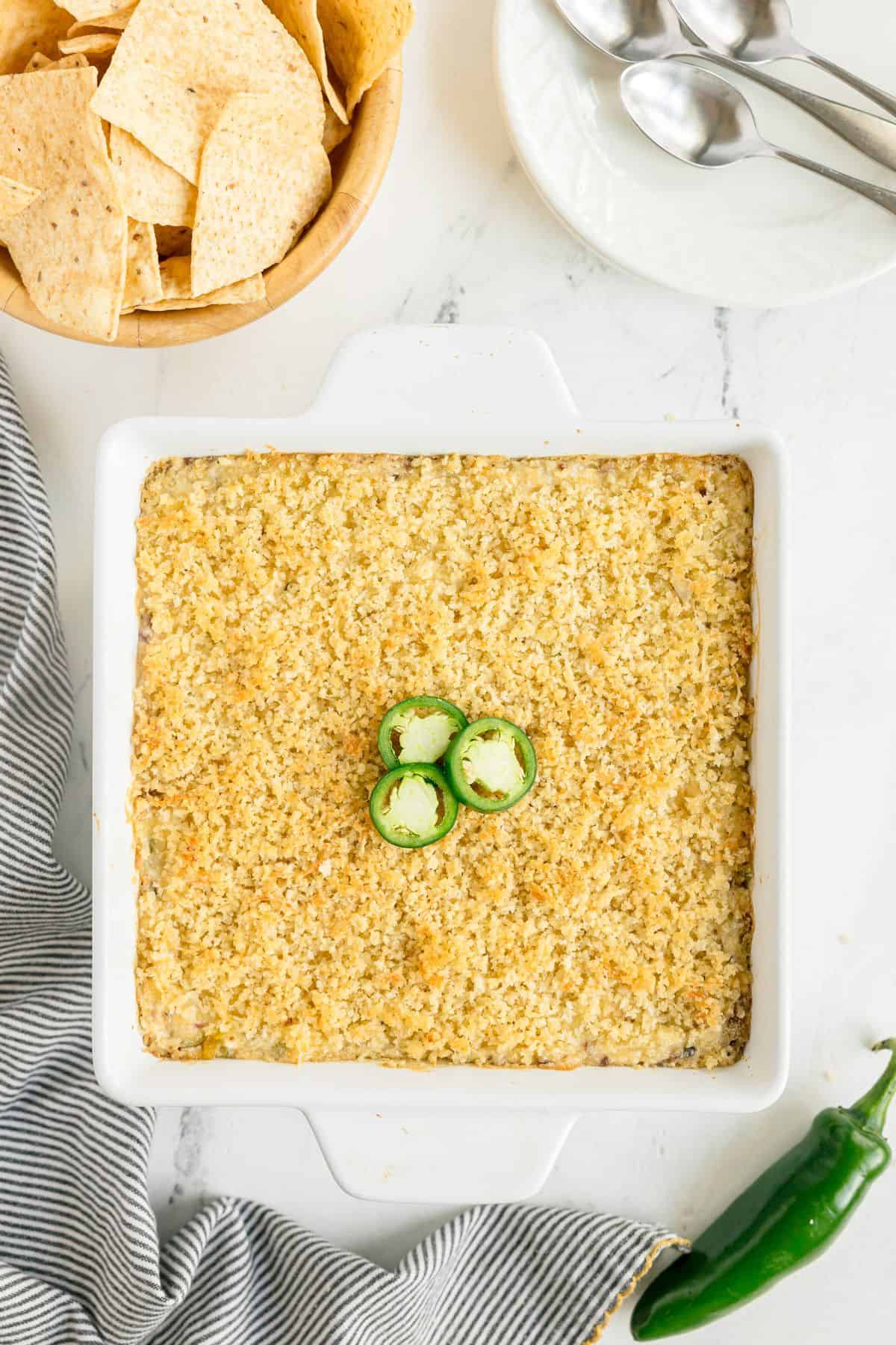 A square white baking dish filled with a crumb-topped casserole, garnished with sliced jalapeños, next to a bowl of tortilla chips, plates, and spoons.