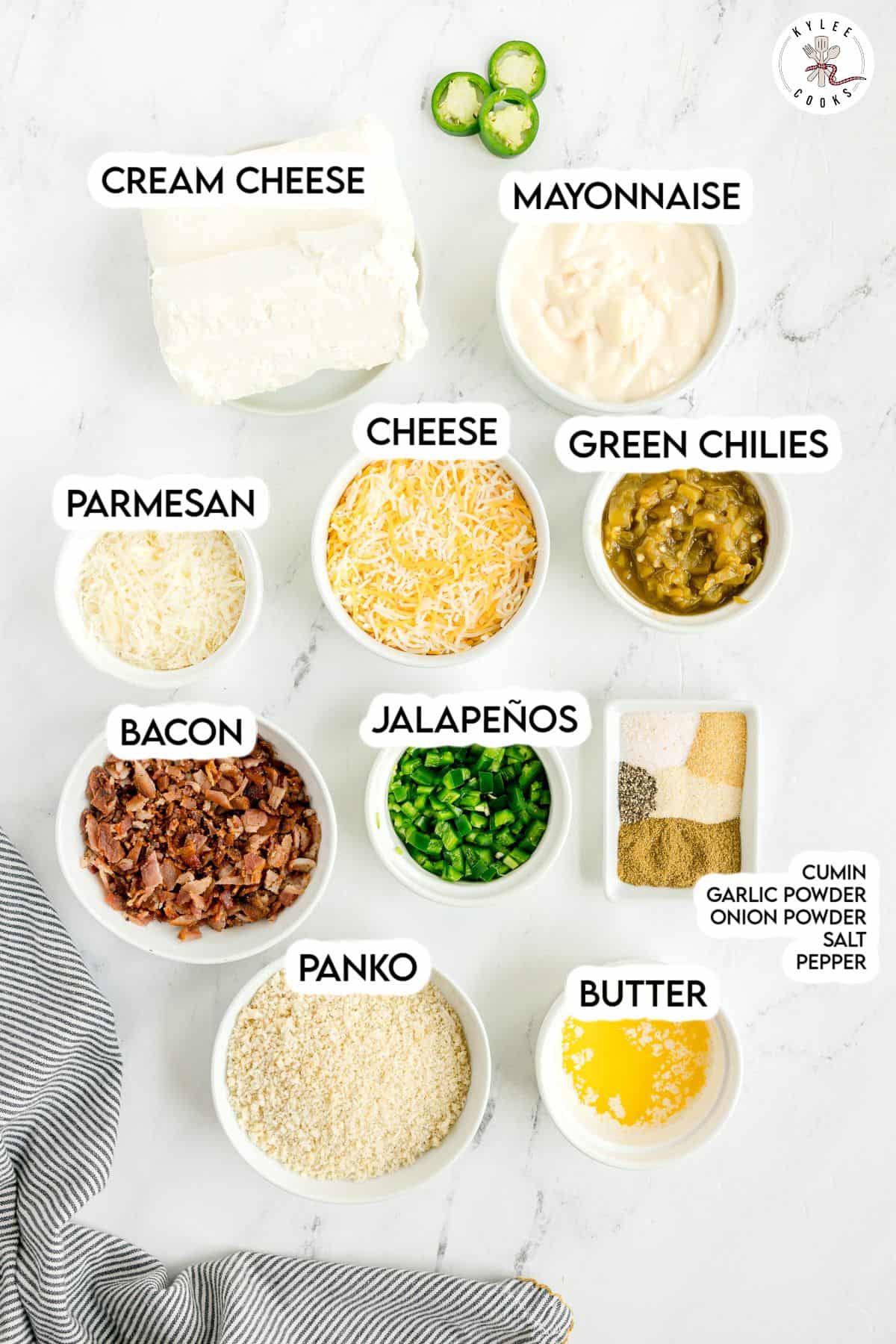 Nine labeled bowls contain cream cheese, mayonnaise, shredded cheese, green chilies, parmesan, bacon, chopped jalapeños, panko, melted butter, and spices on a white surface.