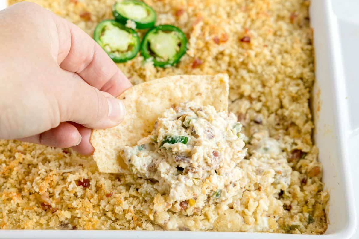 A hand scooping a creamy, jalapeño-topped dip with a tortilla chip from a breadcrumb-topped casserole dish.
