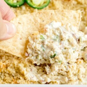 A hand holds a tortilla chip scooping a creamy, cheesy dip with visible herbs and crumb topping, with sliced jalapeños and more dip in the background.