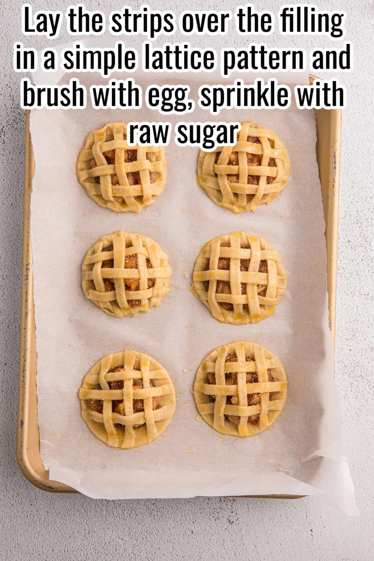 Six unbaked mini pies with lattice crusts are arranged on a parchment-lined baking sheet, sprinkled with raw sugar, ready for baking.