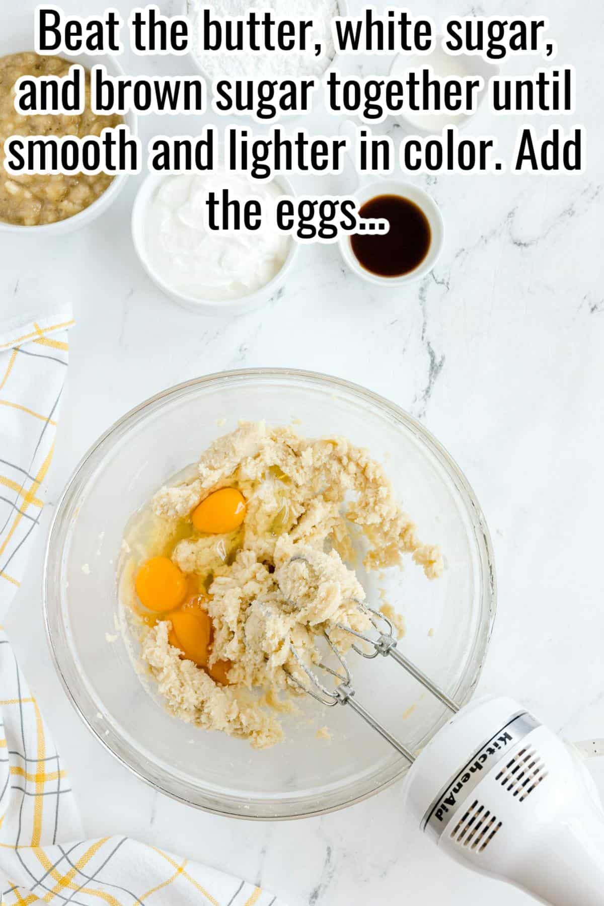 A mixing bowl with creamed butter, white sugar, brown sugar, and three eggs, with a hand mixer and various baking ingredients nearby on a white countertop.