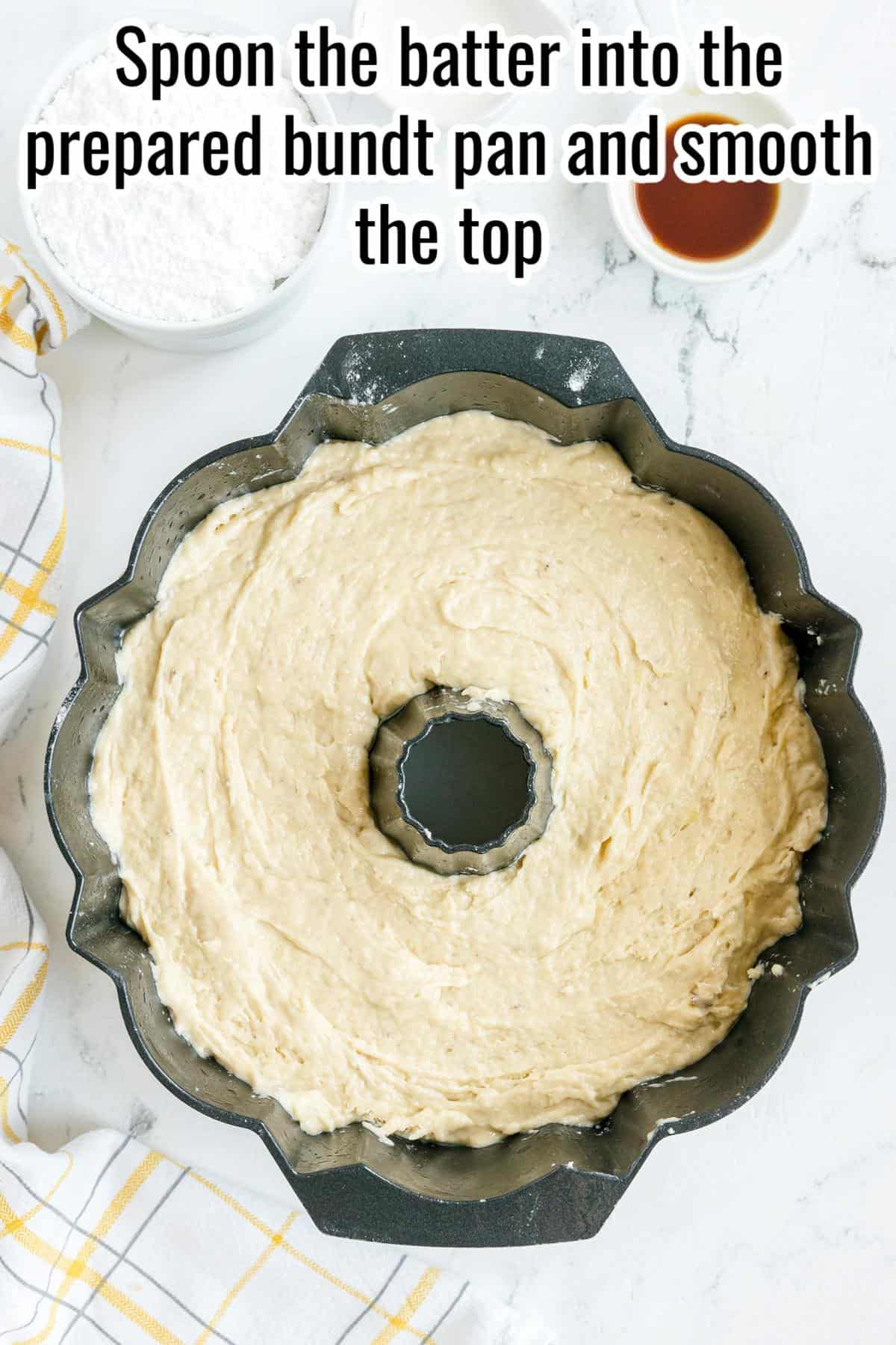 Bundt pan filled with batter, ready to bake. A bowl of flour, cup of liquid, and a yellow-checkered towel are nearby on a marble surface.