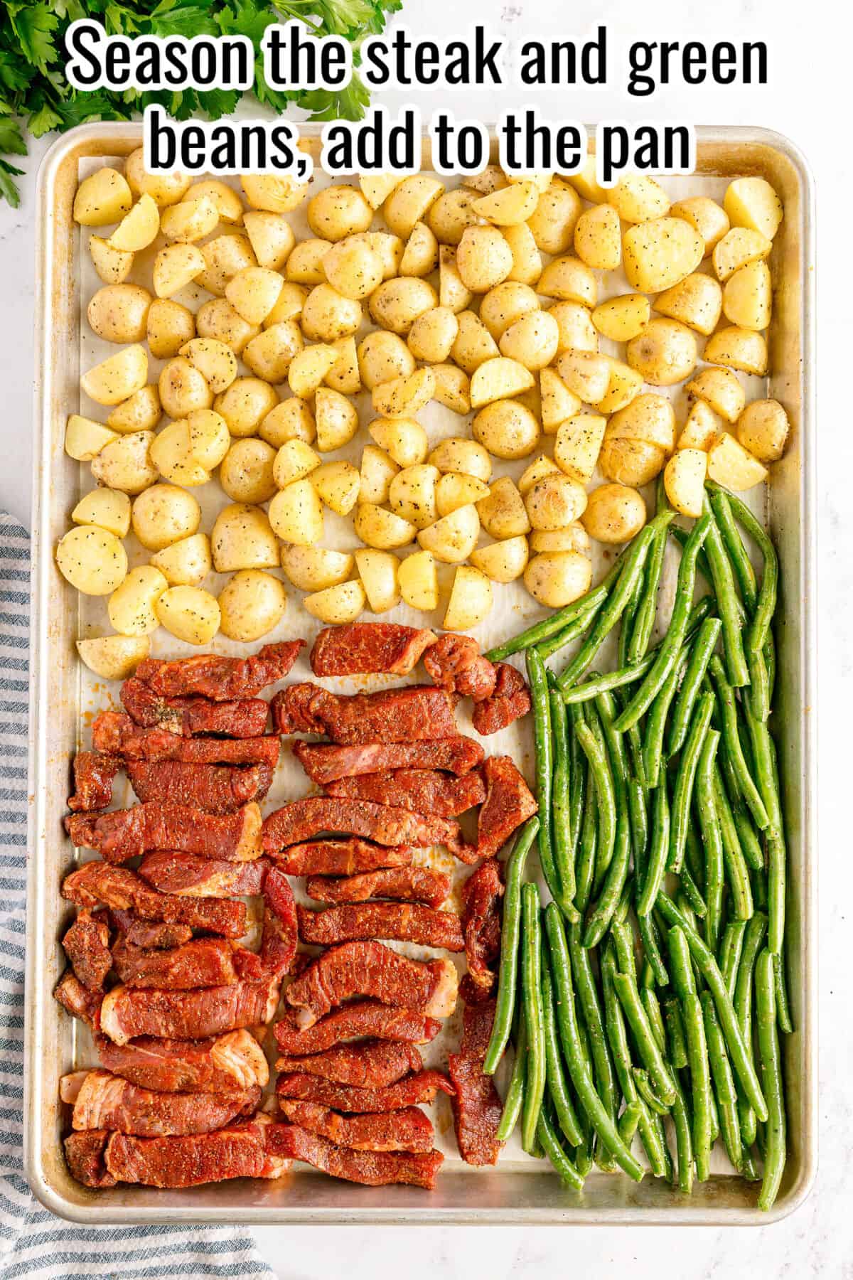 A sheet pan with seasoned potato pieces, raw steak strips with spices, and fresh green beans arranged in sections, ready to bake.