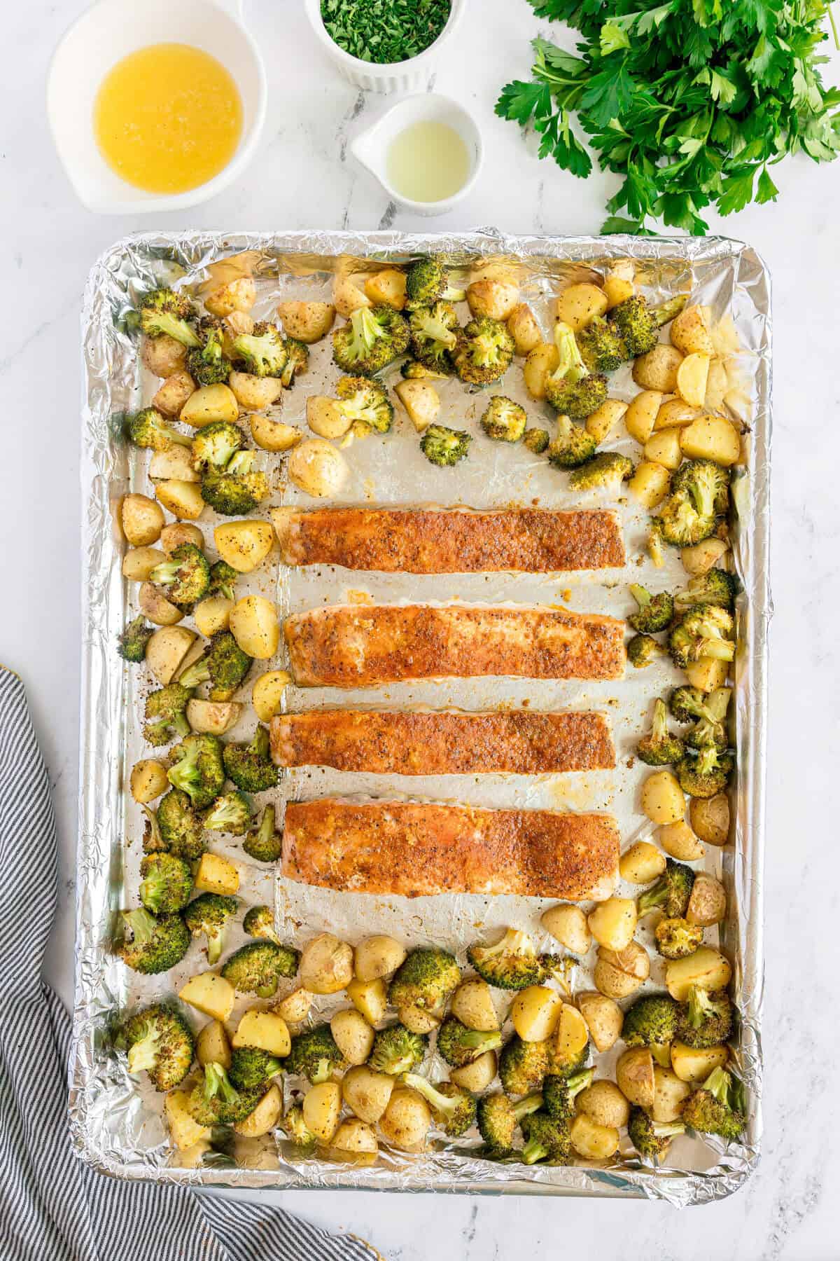 Sheet pan with four seasoned salmon fillets, roasted broccoli, and diced potatoes on foil, surrounded by bowls of sauce, chopped herbs, and fresh parsley.
