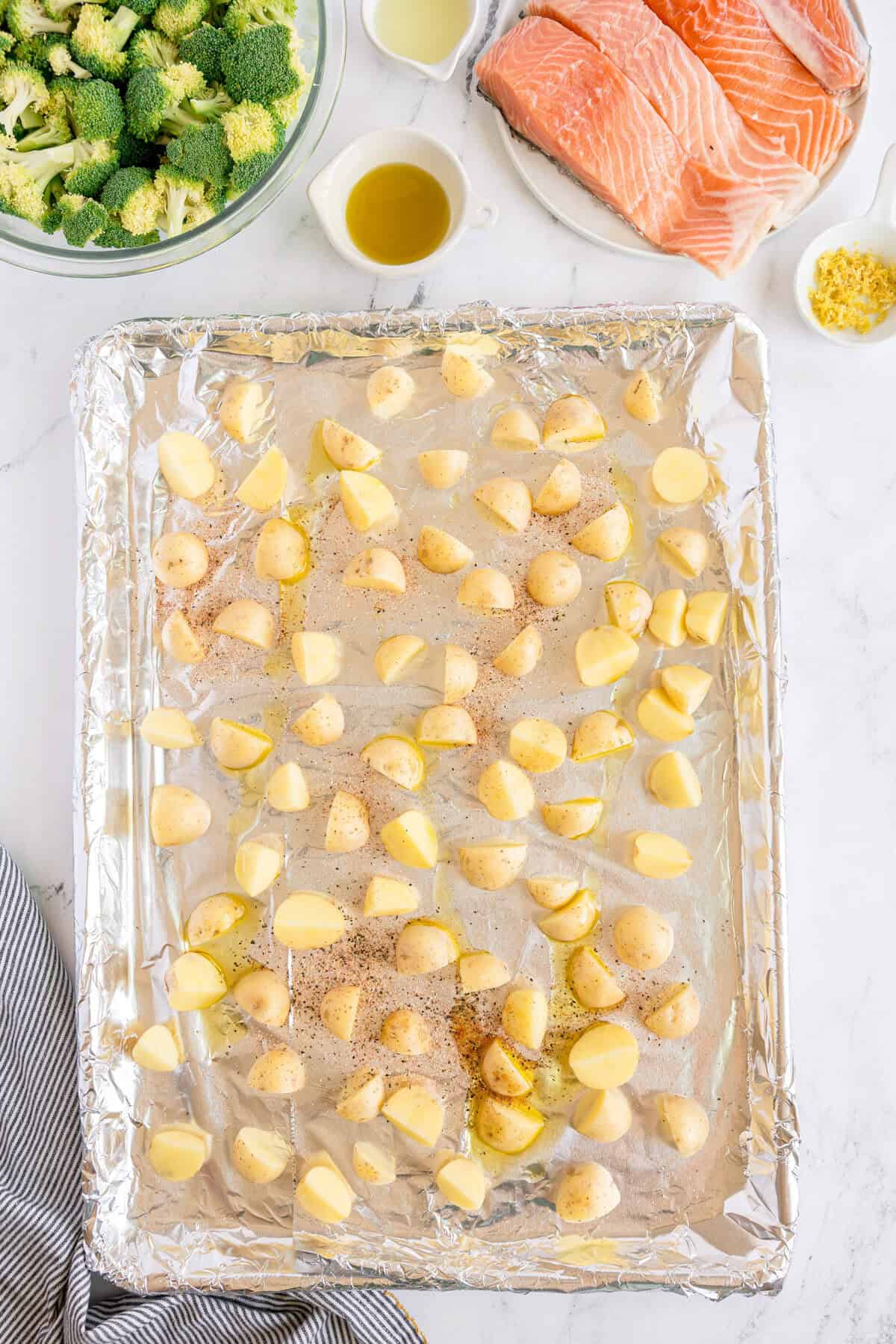 A baking sheet with seasoned potato chunks on foil, next to bowls of broccoli, olive oil, minced garlic, and raw salmon fillets on a white surface.