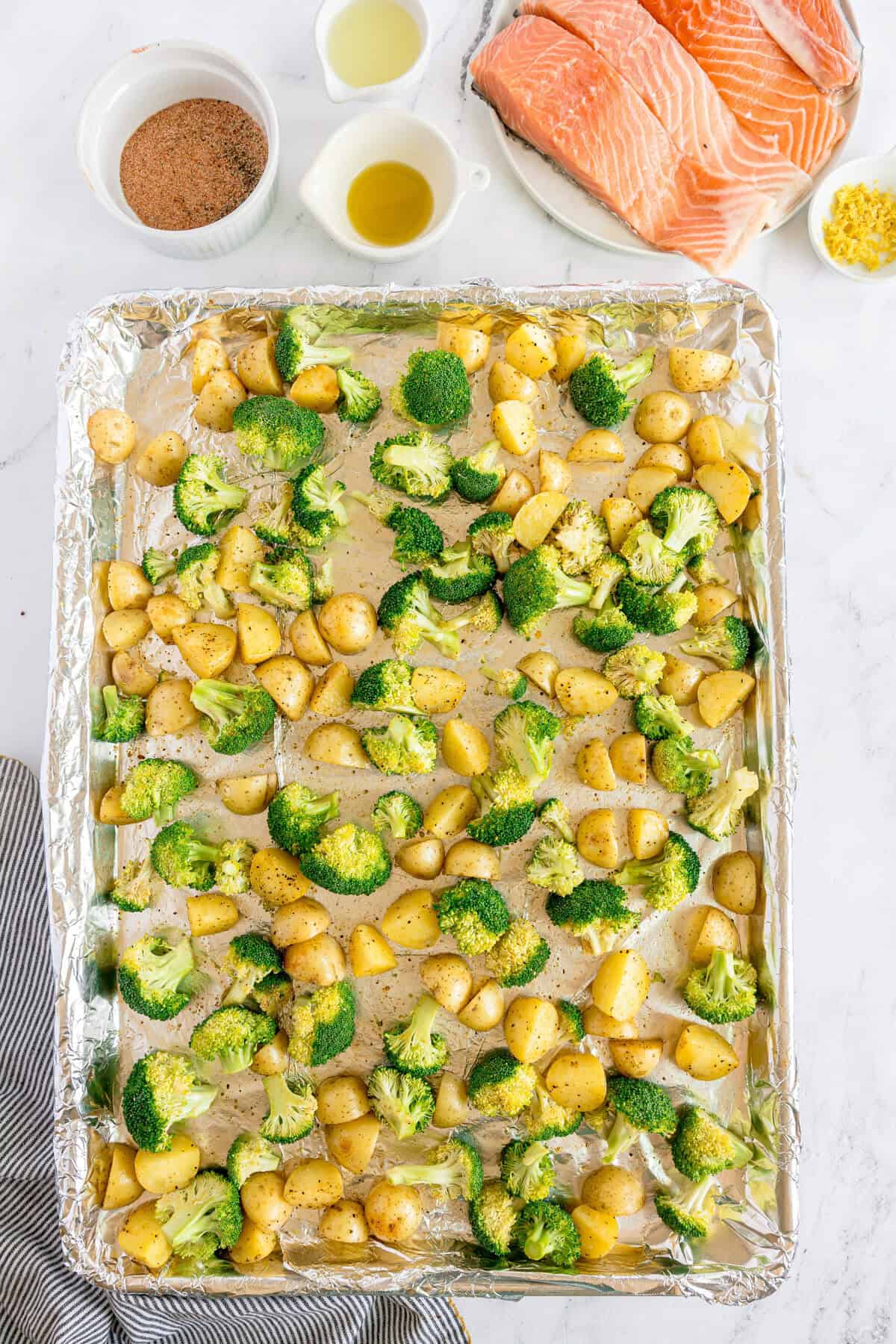 A foil-lined baking sheet with raw broccoli florets and diced potatoes, next to uncooked salmon fillets and small bowls of oil, seasoning, and lemon zest on a white surface.