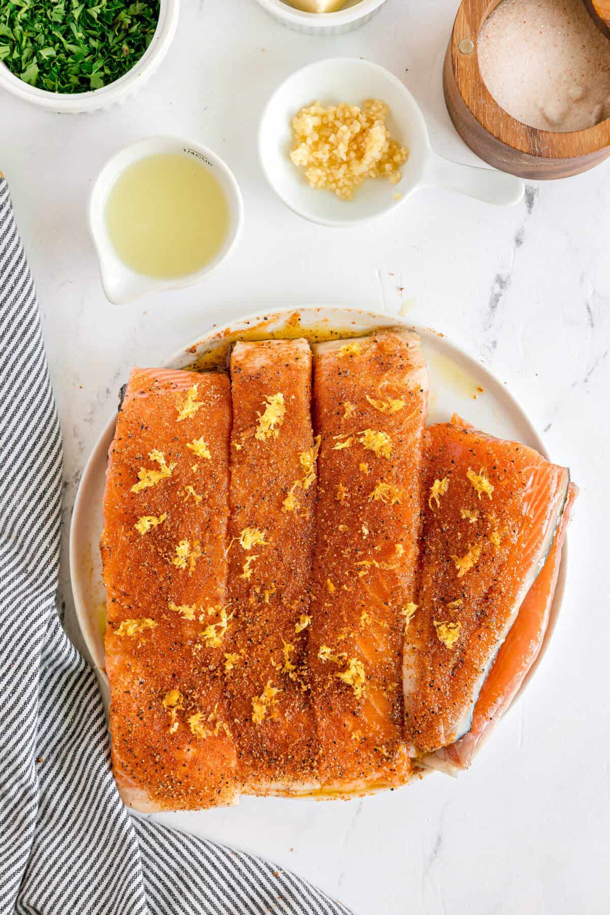 A plate of raw salmon fillets seasoned with spices and lemon zest sits on a white surface next to small bowls of chopped herbs, minced garlic, lemon juice, and salt.