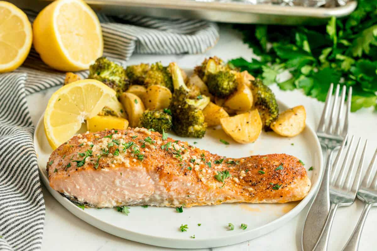 A plate with baked salmon, roasted broccoli, and potatoes, garnished with herbs and served with lemon slices. Forks and chopped parsley are nearby.