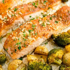 Baked salmon fillets topped with herbs and minced garlic, served with roasted broccoli and potatoes on a sheet pan.