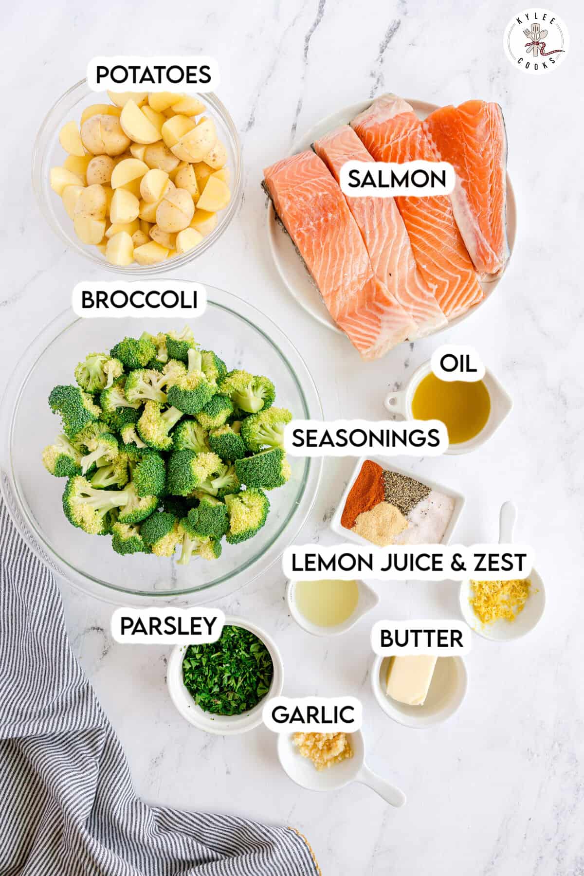 Ingredients for a sheet pan salmon dish laid out on a white surface, including salmon fillets, potatoes, broccoli, seasonings, oil, butter, garlic, lemon juice and zest, and chopped parsley.