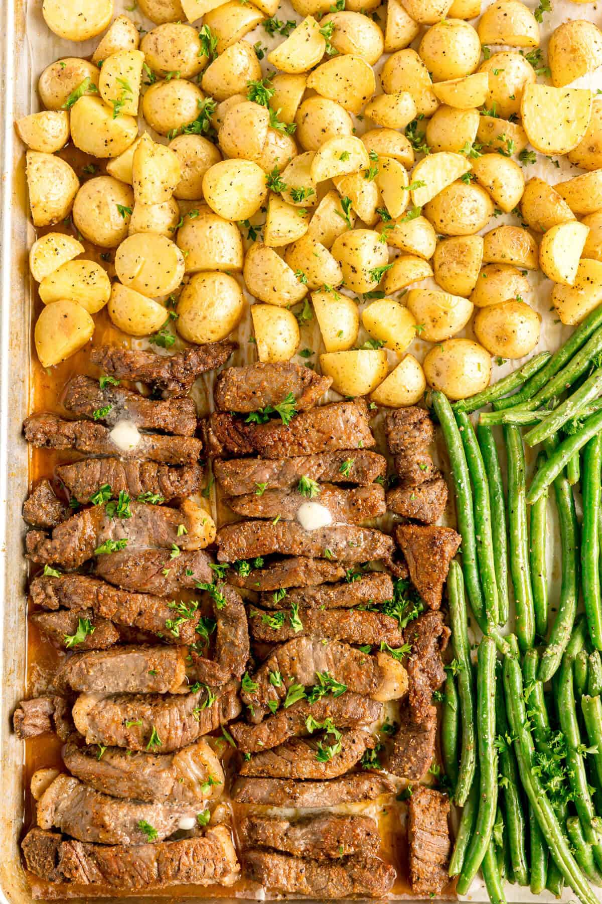 Sheet pan with roasted baby potatoes, sliced steak topped with herbs and butter, and green beans arranged in separate sections.