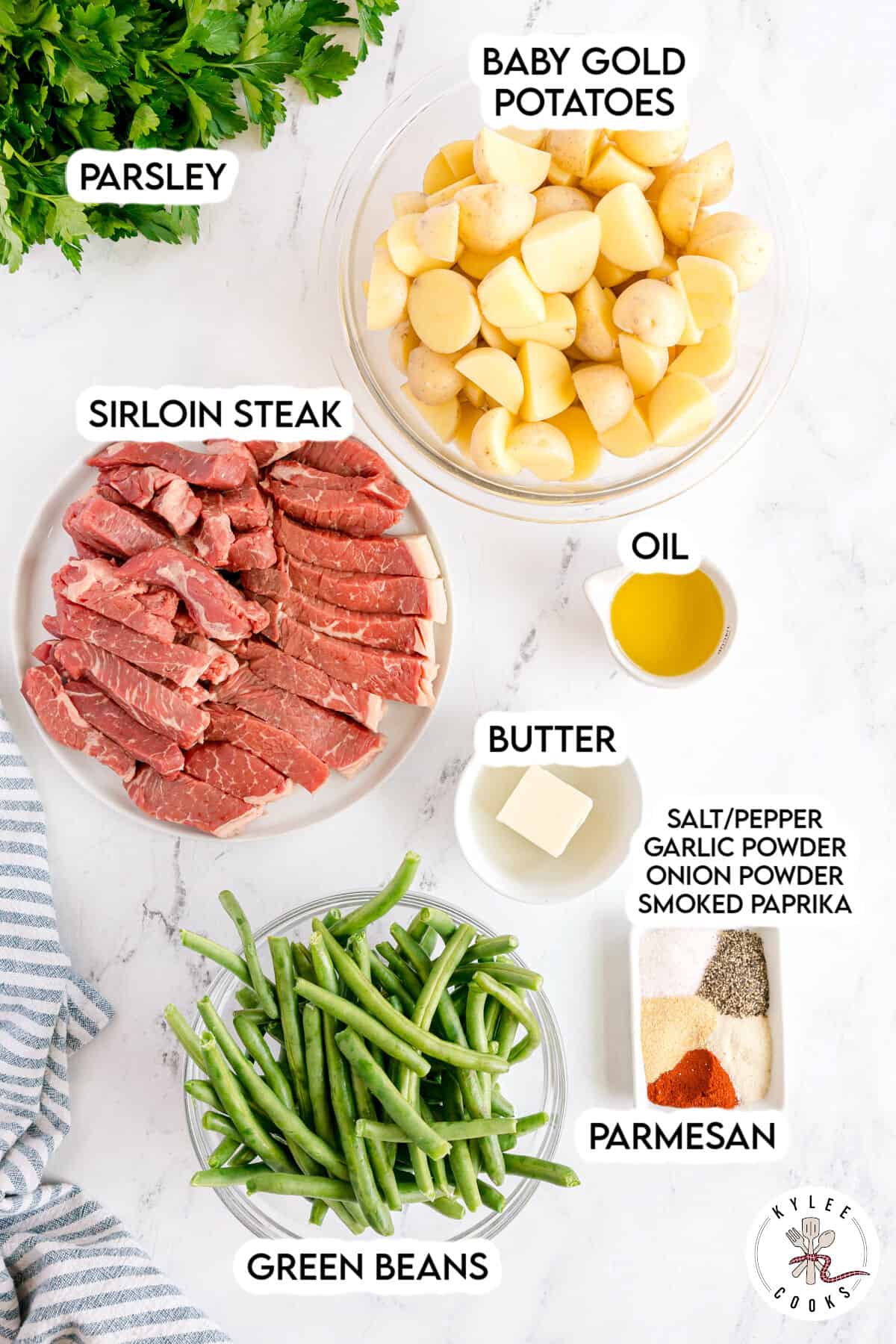 Bowls with labeled ingredients: sliced sirloin steak, baby gold potatoes, green beans, parsley, oil, butter, and assorted spices on a white surface.