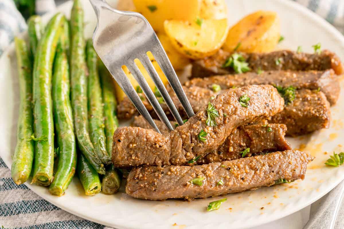 A fork holds a piece of seasoned steak on a plate with green beans and roasted potatoes.