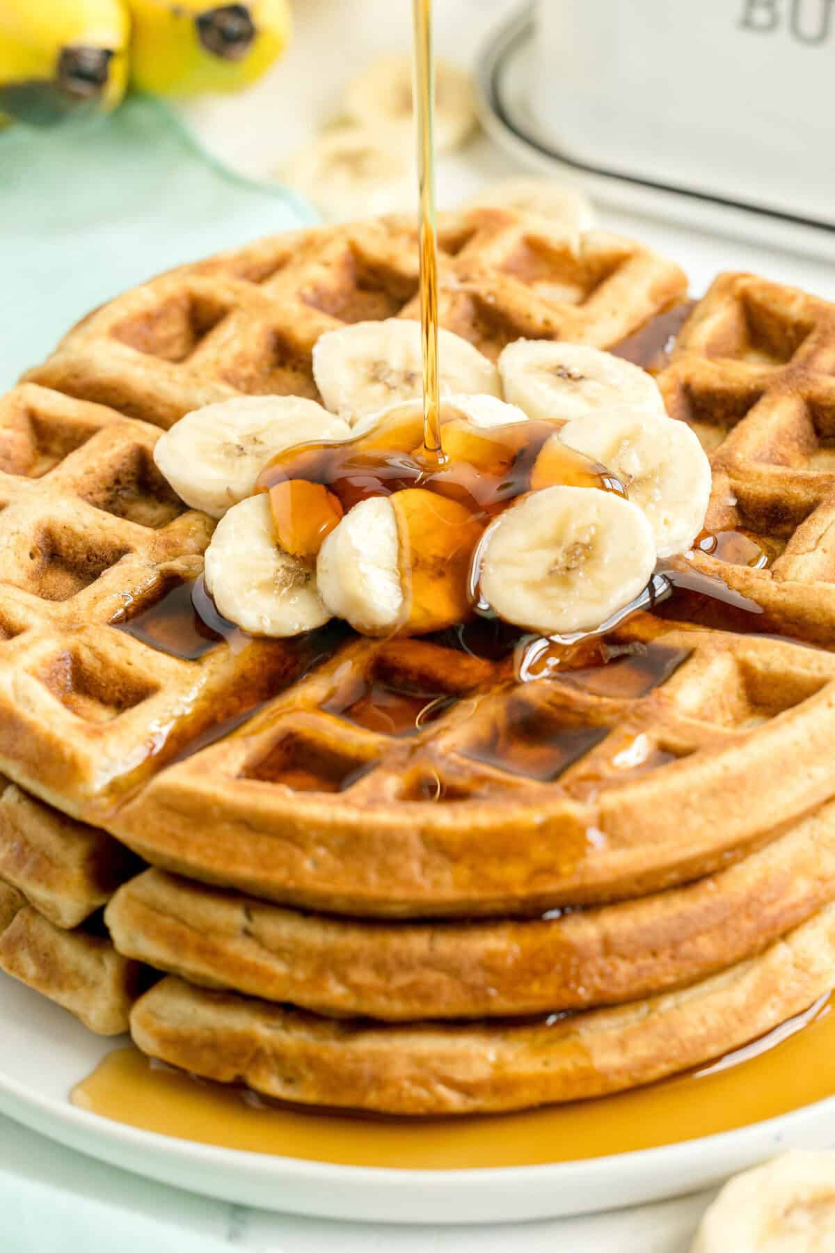 A stack of waffles topped with banana slices is being drizzled with syrup on a white plate.