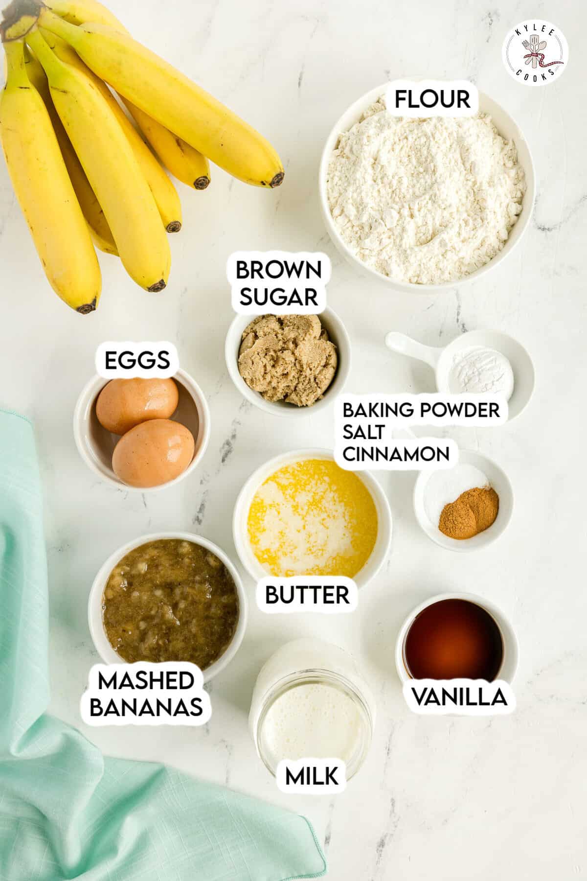 Top-down view of ingredients for banana bread, including bananas, flour, brown sugar, eggs, baking powder, cinnamon, butter, vanilla, mashed bananas, and milk on a white surface.