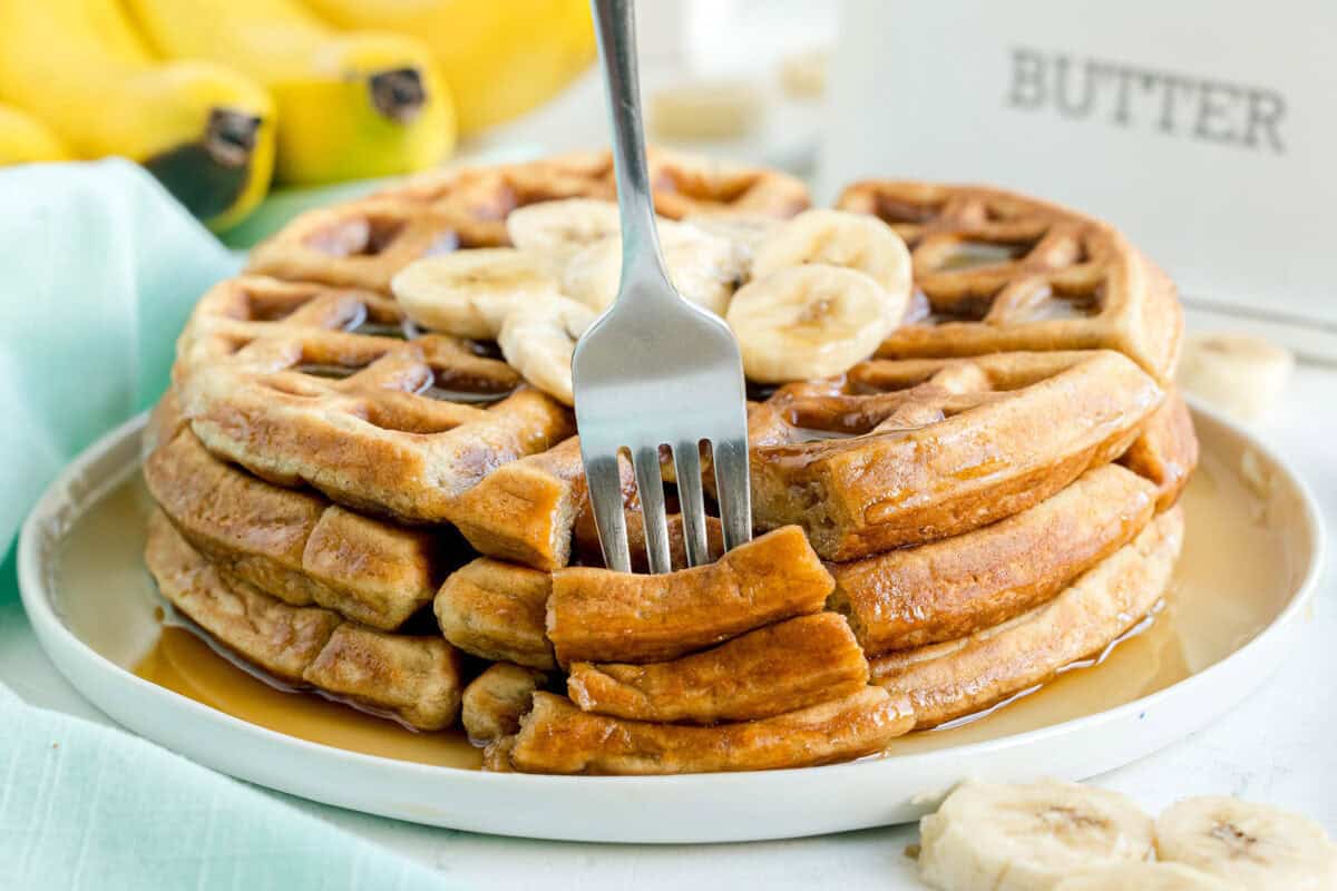 A fork slices into a stack of waffles topped with banana slices and syrup on a white plate; bananas and a butter dish are in the background.