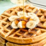 Stack of waffles topped with banana slices and syrup being poured over them.