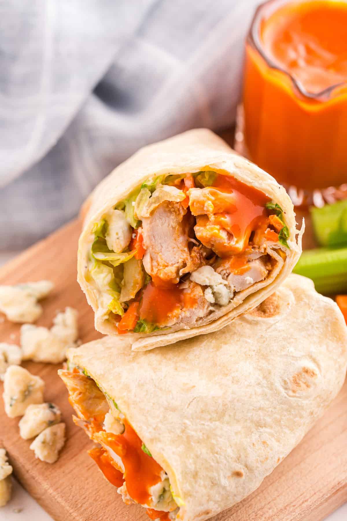 A close-up of Buffalo Chicken Wraps filled with chicken, lettuce, blue cheese crumbles, and orange sauce, placed on a wooden board next to a glass of orange drink.