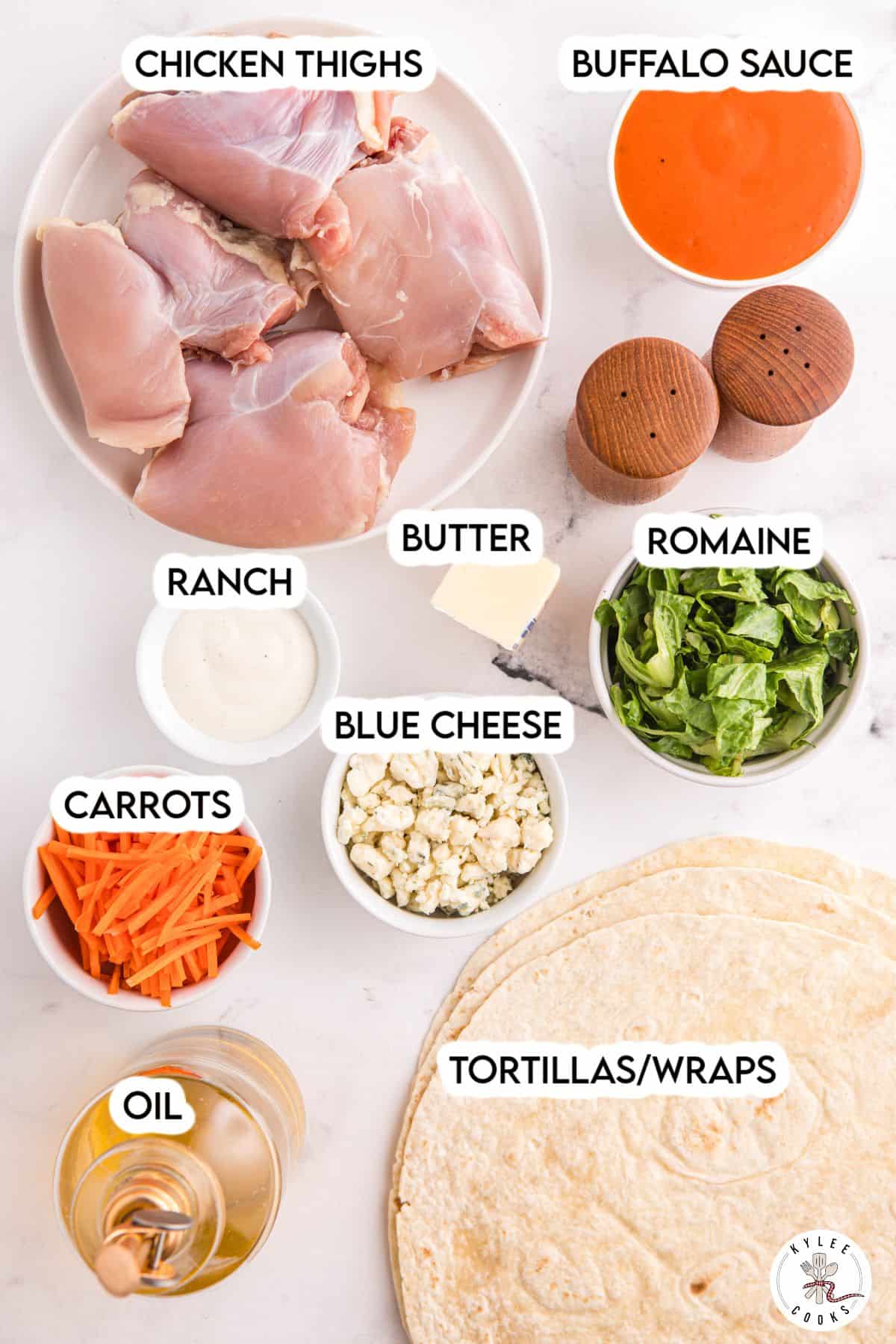 Ingredients for buffalo chicken wraps, including chicken thighs, buffalo sauce, butter, ranch, blue cheese, romaine, carrots, oil, and tortillas displayed on a white surface.