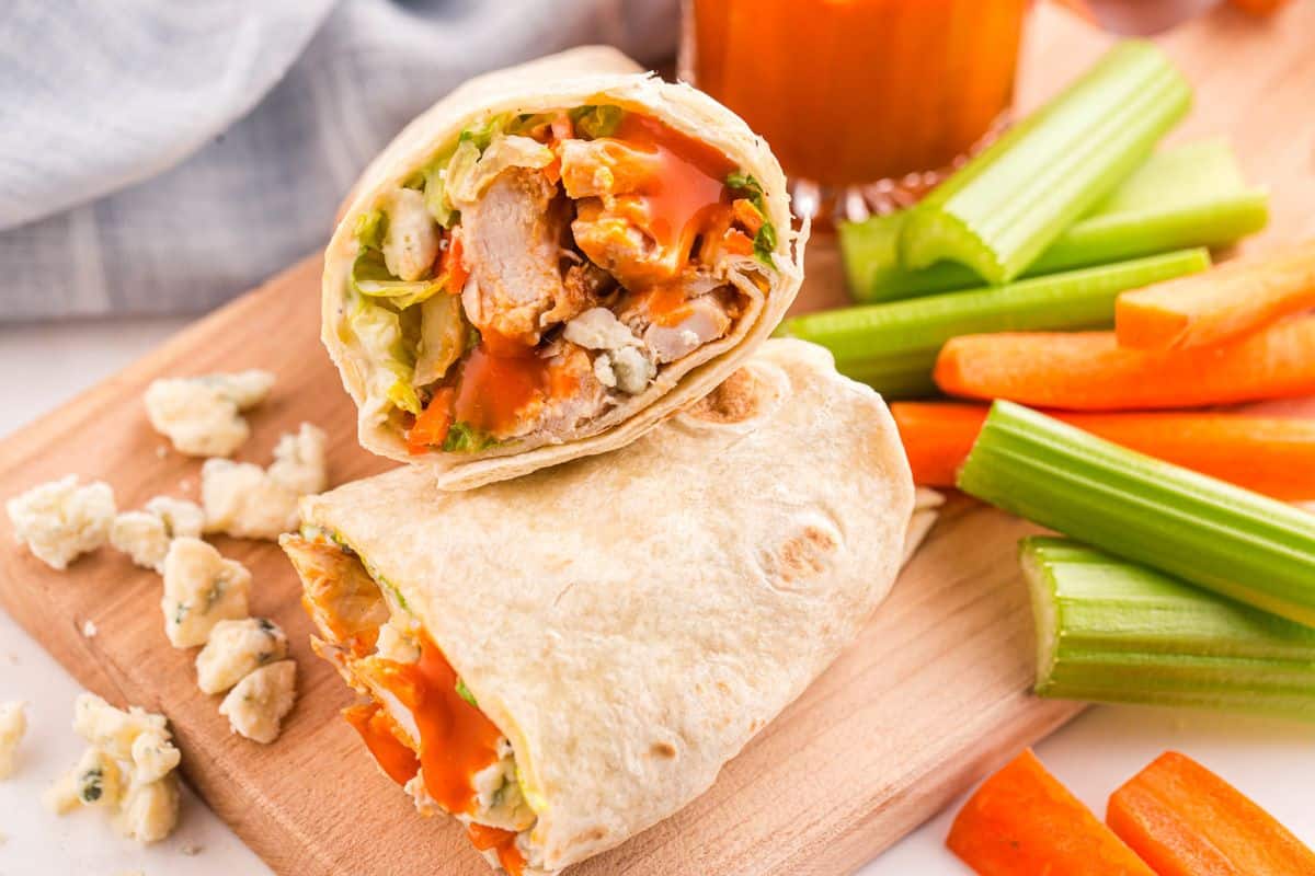 A buffalo chicken wrap cut in half on a wooden board, surrounded by celery sticks, carrot sticks, and blue cheese crumbles.