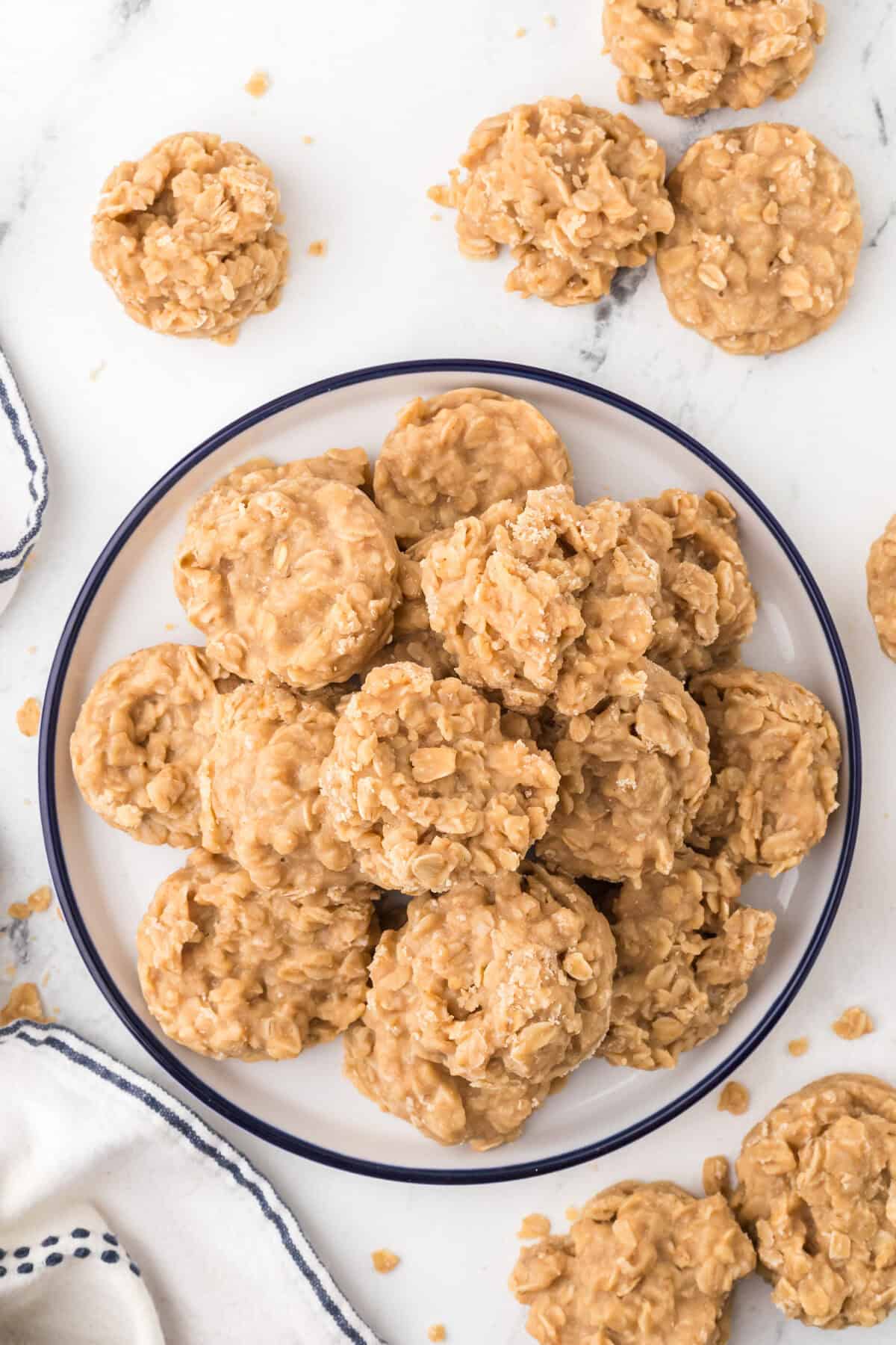 A plate of no-bake peanut butter cookies is surrounded by a few cookies on a white marble surface, next to a white napkin with blue trim.